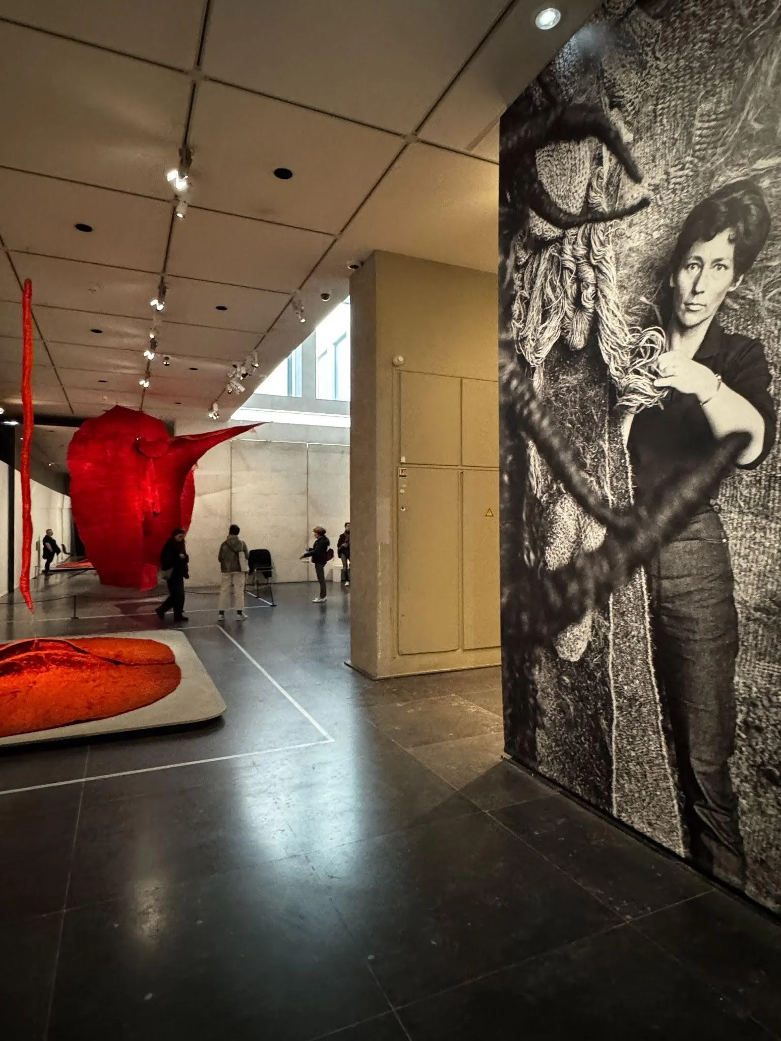 A gallery with a monumental black-and-white portrait of a woman artist holding skeins of fiber, beside suspended and floor-set red woven textile sculptures.