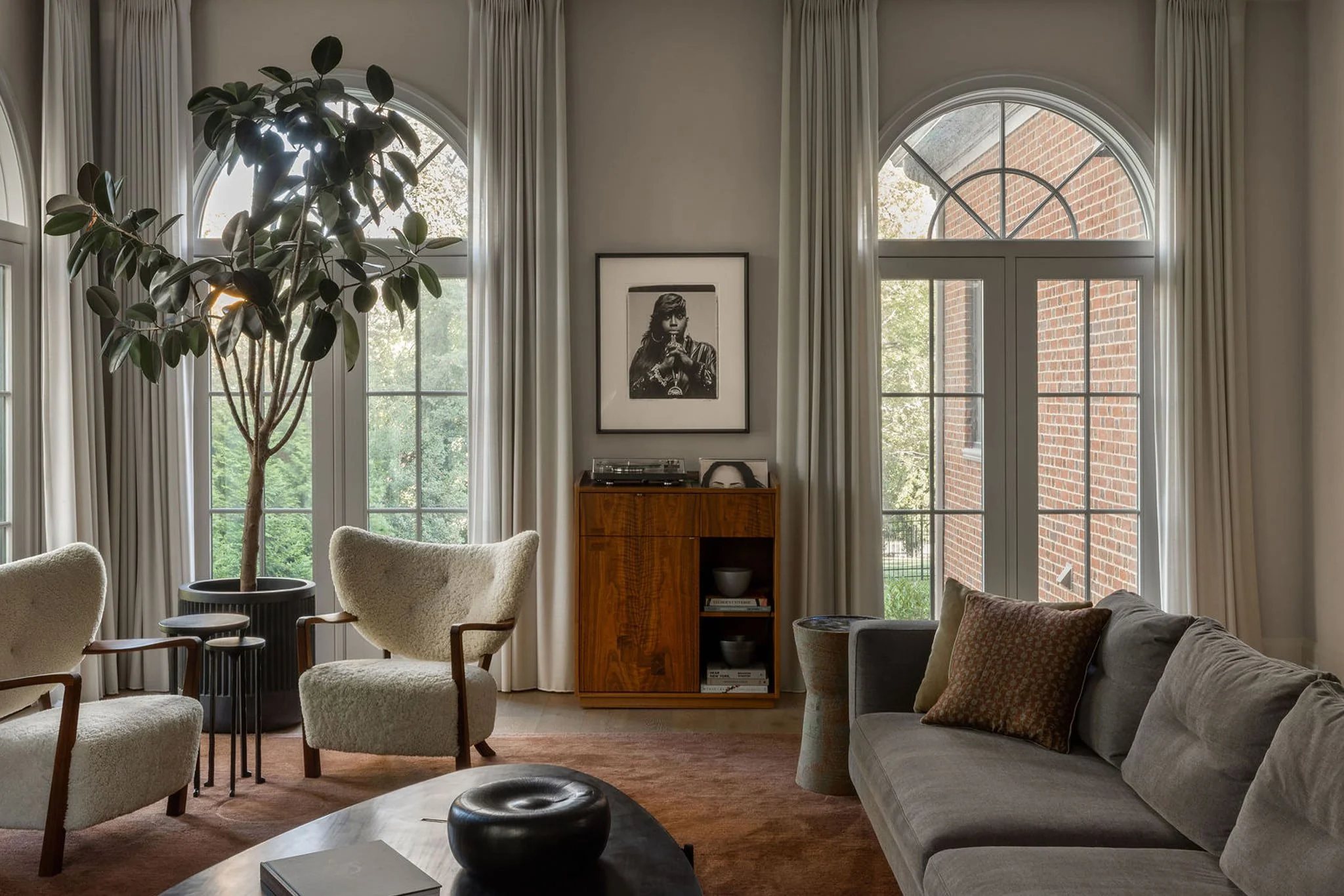 A sunlit living room with tall arched windows, sculptural lounge chairs, and a large fiddle-leaf plant.