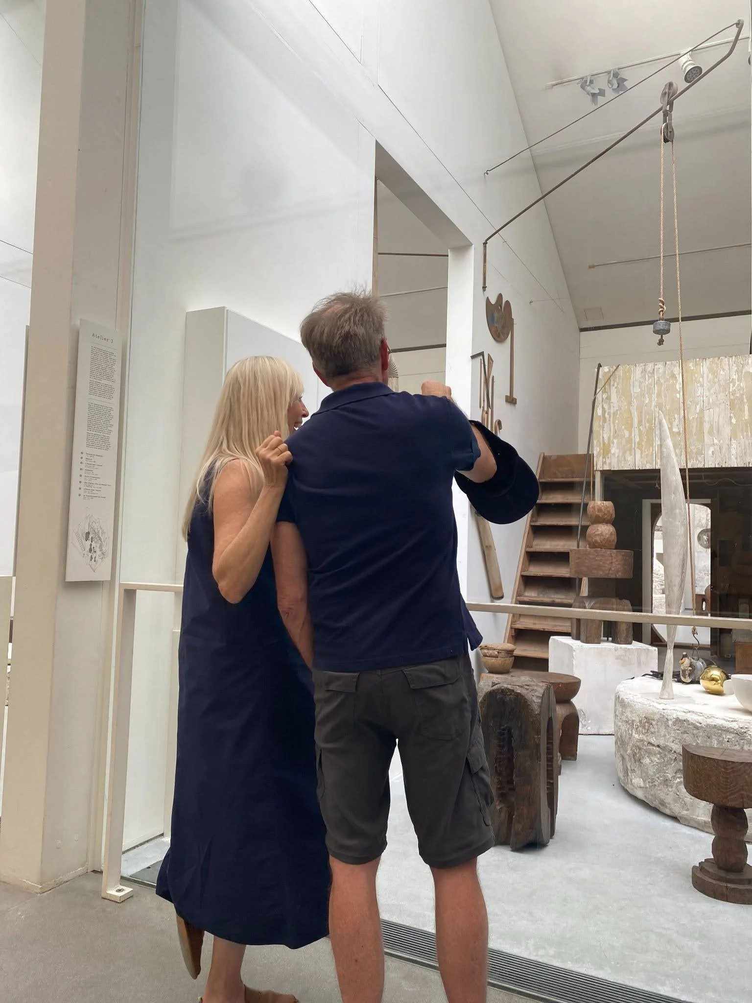 Two visitors on a mezzanine overlooking a sculptor's atelier filled with stacked carved-wood totem sculptures, stone plinths, and a rustic wooden staircase.
