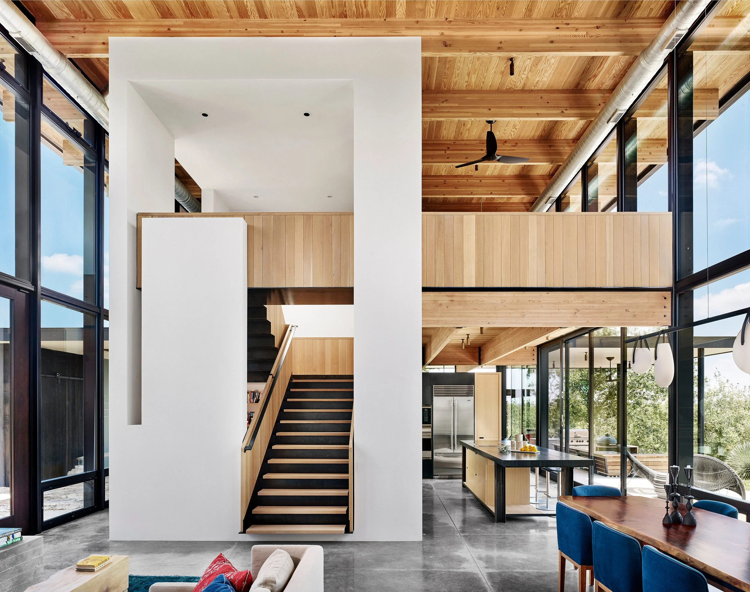 Interior of a modern Hill Country home, showing the kitchen, dining and stairwell to the second floor with plaster walls, concrete floors, white oak millwork, and expansive glazing.