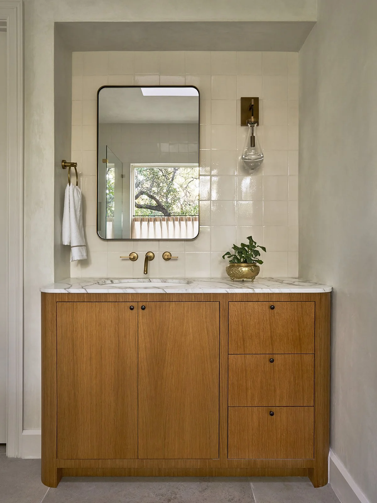 Primary bathroom vanity featuring a custom curved white oak cabinet, marble countertop, metal-framed mirror, and plaster walls.