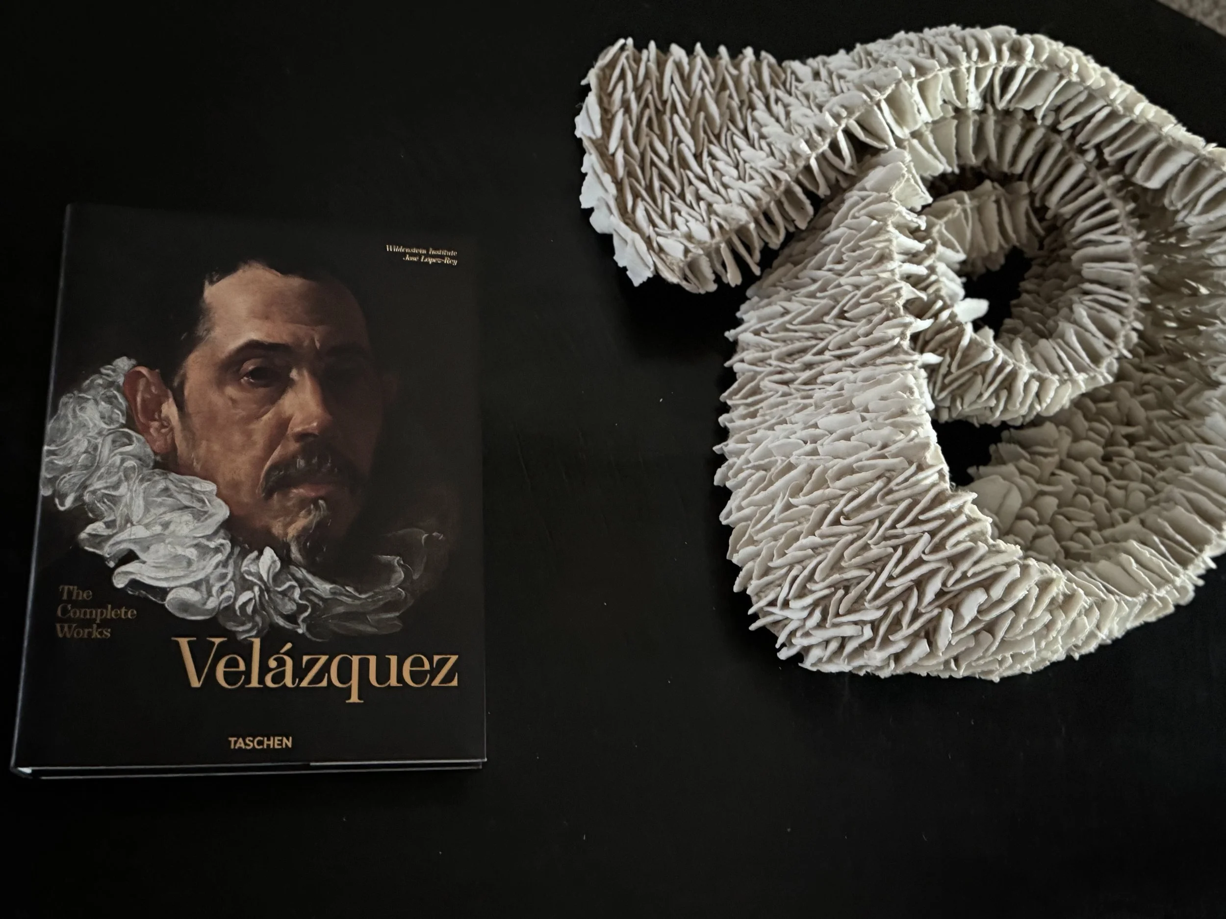 A coiled sculpture of ruffled white porcelain petals beside a Taschen ‘Velázquez: The Complete Works’ monograph on a black surface.