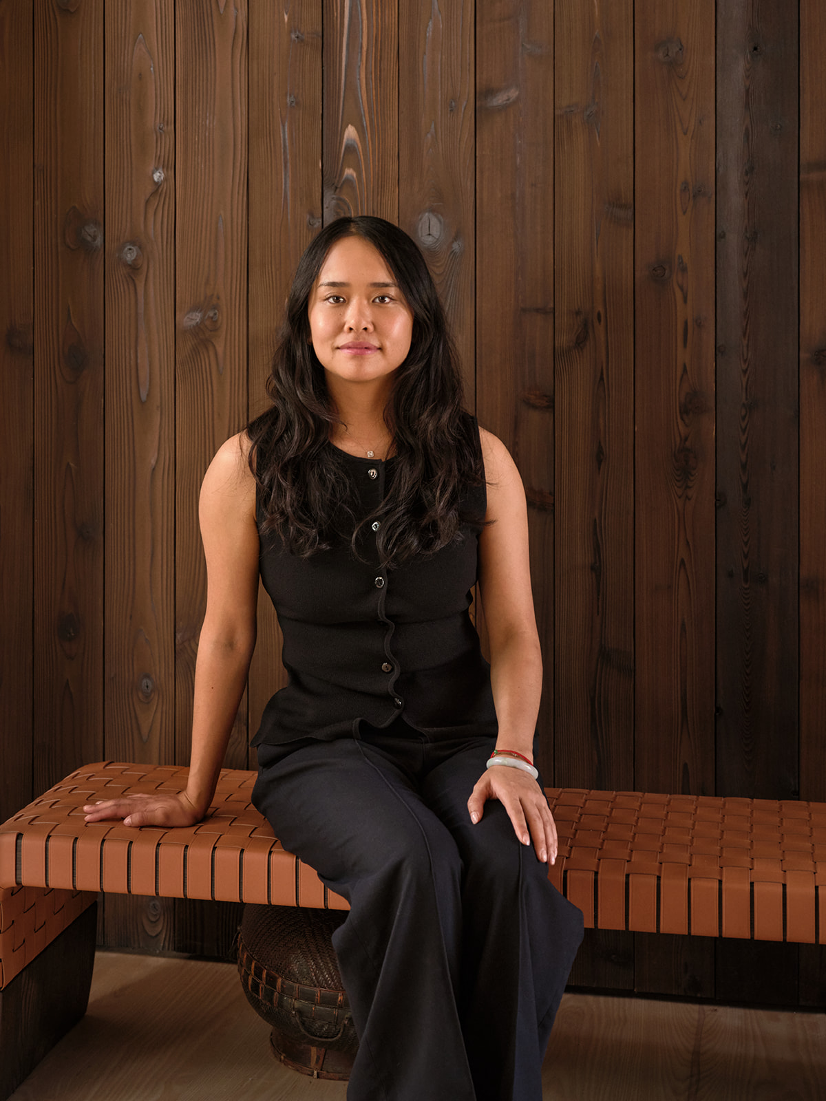 Hil Wong seated on a woven leather-strap bench against a dark vertical wood-plank wall.